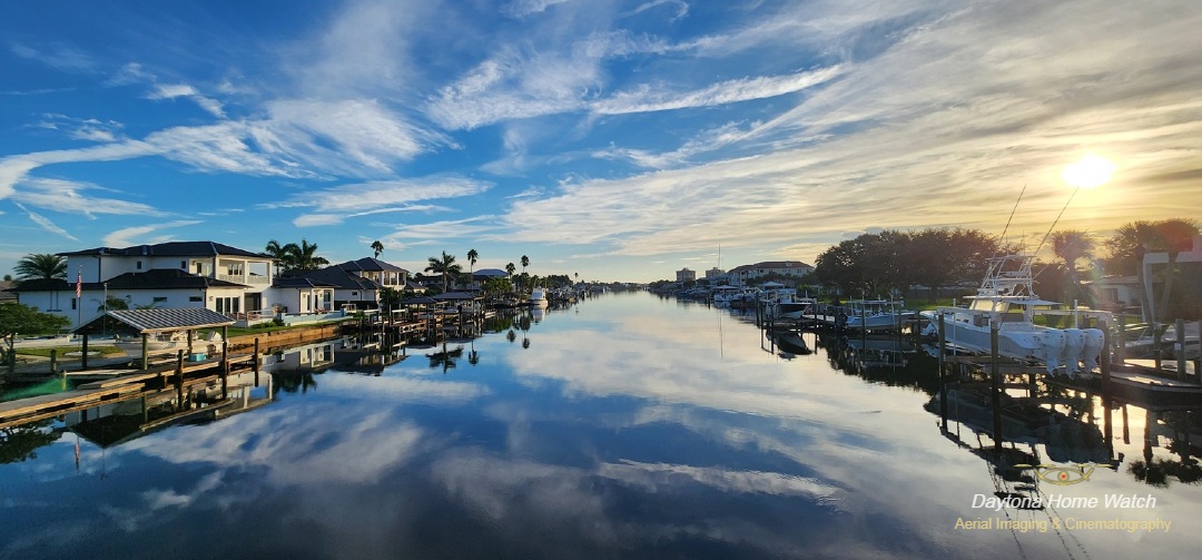 Panoramic aerial photograph overlooking Barracuda Waterway in New Smyrna Beach, Florida, where beautiful waterfront homes, private docks, and boats frame remarkably calm waters reflecting a brilliant blue sky and sweeping cloud formations.